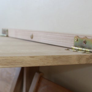 Puede incluir: Una mesa plegable de madera con un acabado de madera clara y bisagras doradas. La mesa est&aacute; unida a la pared con bisagras.