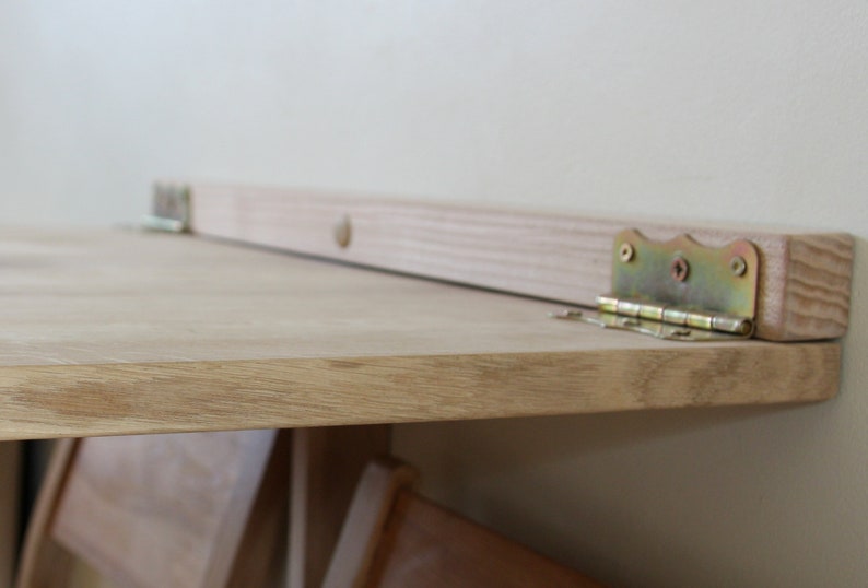 Puede incluir: Una mesa plegable de madera con un acabado de madera clara y bisagras doradas. La mesa est&aacute; unida a la pared con bisagras.