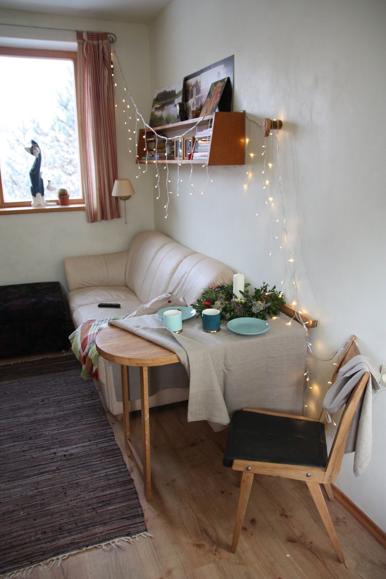 May include: A small, round wooden table with a white tablecloth set for two. There are two blue mugs, a green wreath with candles, and a blue plate on the table. A wooden chair with a black seat sits next to the table. The table is in a living room with a couch and a window in the background.