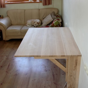 May include: A light wood folding table mounted to a wall. The table is made of solid wood and has a simple design. The table is folded up against the wall and is not in use.
