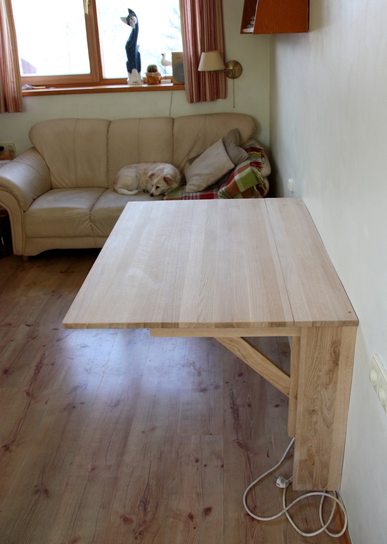 May include: A light wood folding table mounted to a wall. The table is made of solid wood and has a simple design. The table is folded up against the wall and is not in use.