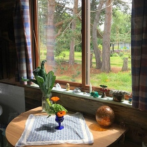 K&ouml;nnte beinhalten: Ein Holztisch mit einer wei&szlig;en Tischdecke mit blauem Muster. Eine Vase mit orangefarbenen Blumen und gr&uuml;nen Bl&auml;ttern steht auf dem Tisch. Eine gro&szlig;e, runde, bernsteinfarbene Glaskugel liegt auf dem Tisch. Ein Fenster mit Blick auf B&auml;ume und einen gr&uuml;nen Rasen befindet sich im Hintergrund.