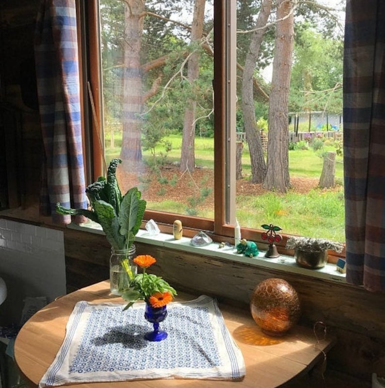 K&ouml;nnte beinhalten: Ein Holztisch mit einer wei&szlig;en Tischdecke mit blauem Muster. Eine Vase mit orangefarbenen Blumen und gr&uuml;nen Bl&auml;ttern steht auf dem Tisch. Eine gro&szlig;e, runde, bernsteinfarbene Glaskugel liegt auf dem Tisch. Ein Fenster mit Blick auf B&auml;ume und einen gr&uuml;nen Rasen befindet sich im Hintergrund.