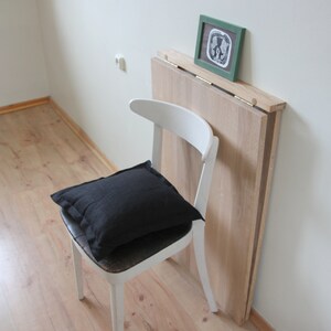 May include: A white wooden chair with a black pillow on the seat. A light wood folding table is leaning against the wall. A small framed picture with a green frame rests on the table.