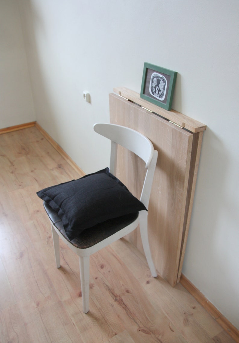 May include: A white wooden chair with a black pillow on the seat. A light wood folding table is leaning against the wall. A small framed picture with a green frame rests on the table.
