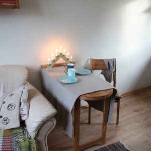 May include: A small table with a gray tablecloth set for two with two blue and white ceramic mugs and two blue plates. The table is next to a white couch with a throw blanket and a pillow. There is a wooden candle holder with lit candles on the table.