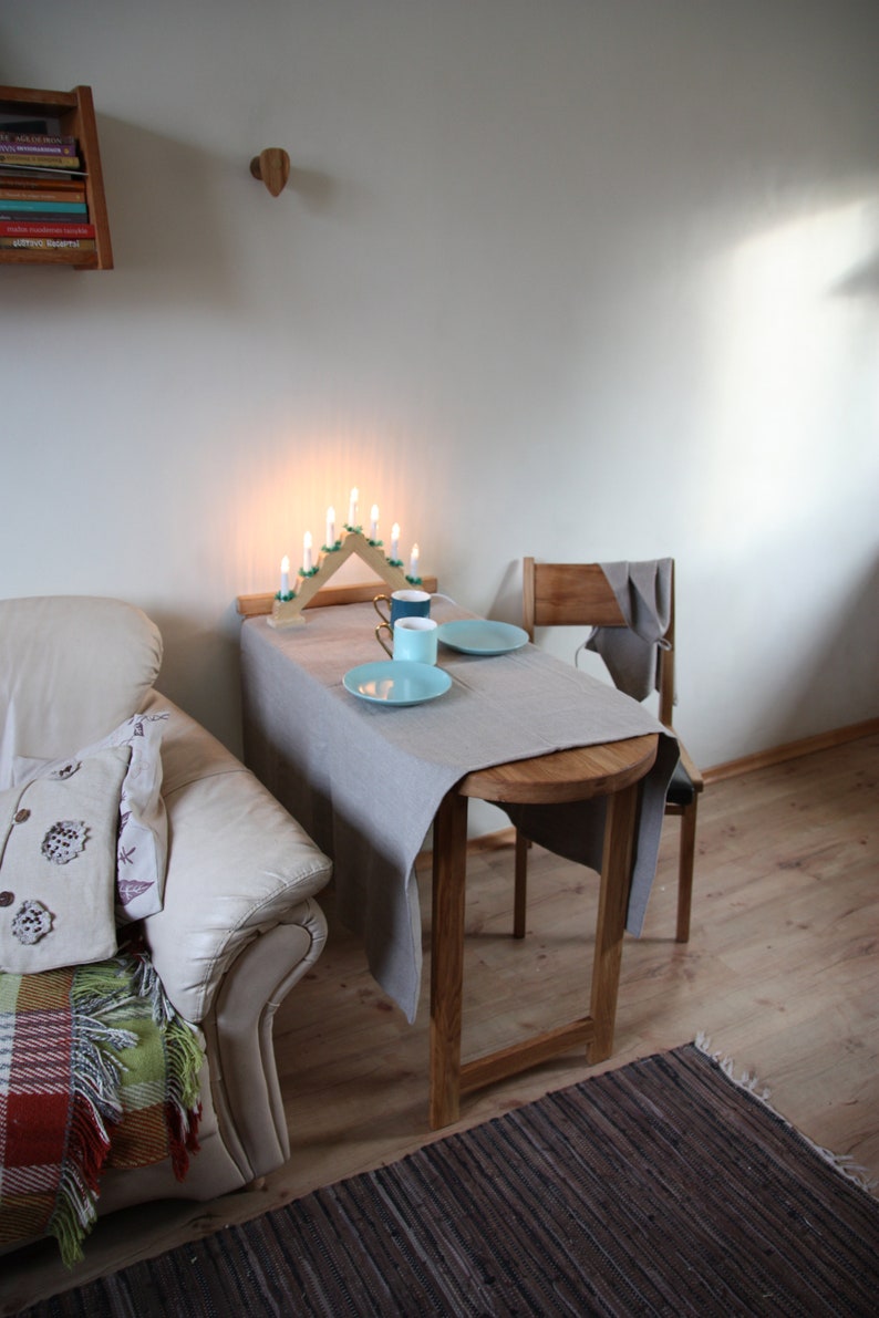 May include: A small table with a gray tablecloth set for two with two blue and white ceramic mugs and two blue plates. The table is next to a white couch with a throw blanket and a pillow. There is a wooden candle holder with lit candles on the table.