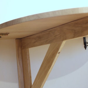 K&ouml;nnte beinhalten: Ein klappbarer Holztisch mit einer abgerundeten Oberseite und schwarzen Metall-Scharnieren. Der Tisch ist mit einer Holzhalterung an der Wand befestigt.