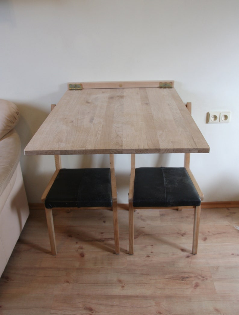 Puede incluir: Una mesa plegable de madera con dos sillas negras. La mesa est&aacute; unida a la pared con bisagras y se puede plegar cuando no se usa.