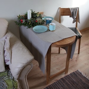 May include: A round wooden table with a gray linen tablecloth. The table is set with two blue and green mugs, a plate, and a candle in a green wreath. The table is next to a white chair with a gray cloth draped over the back.