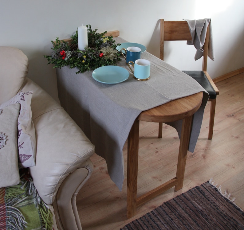 May include: A round wooden table with a gray linen tablecloth. The table is set with two blue and green mugs, a plate, and a candle in a green wreath. The table is next to a white chair with a gray cloth draped over the back.