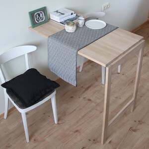May include: A light wood, folding table with a matching chair and a black cushion. A black and white patterned runner is on the table, with a white plate and cup. A framed picture and books are on the table.