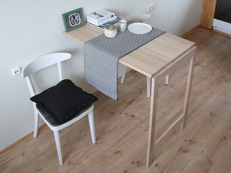 May include: A light wood, folding table with a matching chair and a black cushion. A black and white patterned runner is on the table, with a white plate and cup. A framed picture and books are on the table.
