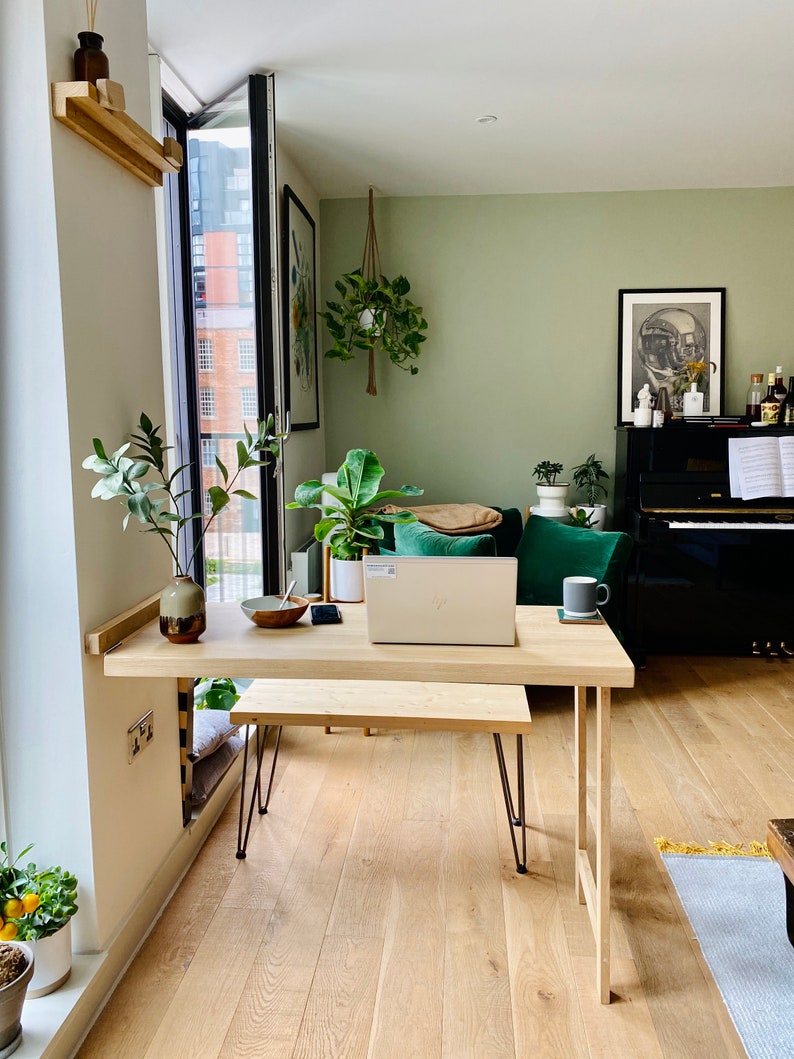 May include: A light wood desk with a laptop, a vase with greenery, and a bowl on top. The desk has black hairpin legs and is set against a white wall. The desk is in a room with a green wall, a black piano, and a green sofa.