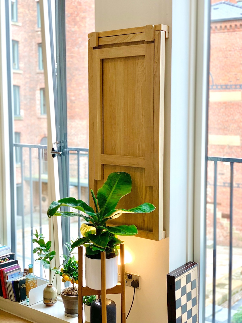 May include: A light wood folding table mounted to a wall, with a plant in a white pot on a wooden stand in front of it.