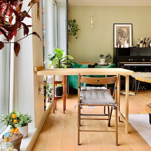 May include: A wooden table with a green chair and a gray cushion. The table is next to a window with a potted plant. The floor is made of wood.