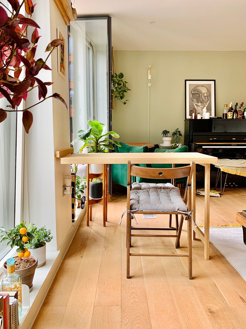 May include: A wooden table with a green chair and a gray cushion. The table is next to a window with a potted plant. The floor is made of wood.
