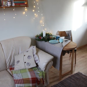 May include: A white leather couch with a plaid throw blanket and a decorative pillow. A small wooden table with a gray tablecloth is set with a plate, cups, and a small green plant.