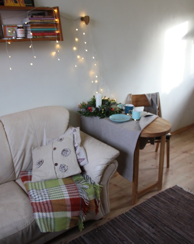 May include: A white leather couch with a plaid throw blanket and a decorative pillow. A small wooden table with a gray tablecloth is set with a plate, cups, and a small green plant.