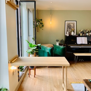 May include: A light wood folding desk with a white wall and a window behind it. The desk is in a living room with green chairs and a black piano.
