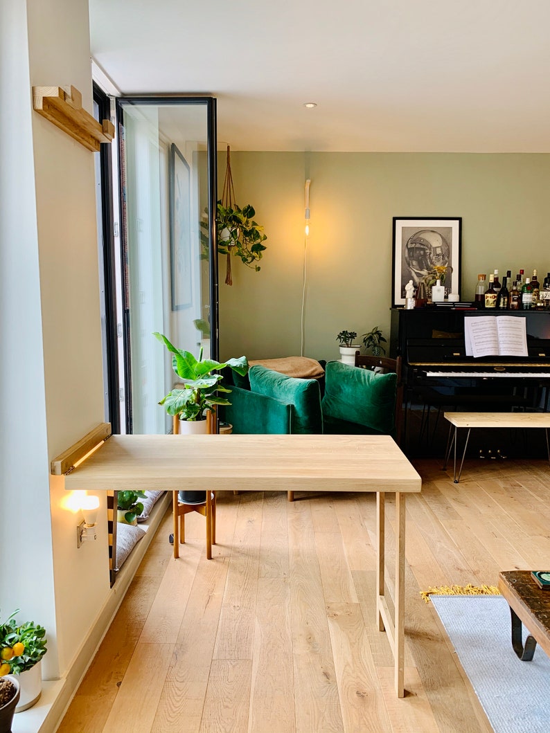 May include: A light wood folding desk with a white wall and a window behind it. The desk is in a living room with green chairs and a black piano.