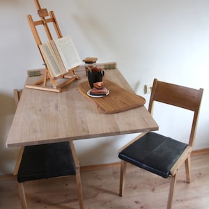 Puede incluir: Una mesa plegable de madera clara con una silla de cuero negro. La mesa tiene un caballete de madera con un libro encima y una taza y un platillo en una bandeja de servicio de madera.