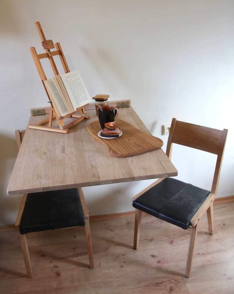 Puede incluir: Una mesa plegable de madera clara con una silla de cuero negro. La mesa tiene un caballete de madera con un libro encima y una taza y un platillo en una bandeja de servicio de madera.