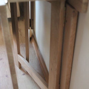 May include: A wooden folding table with a white top and a natural wood finish. The table is folded up and leaning against a wall.