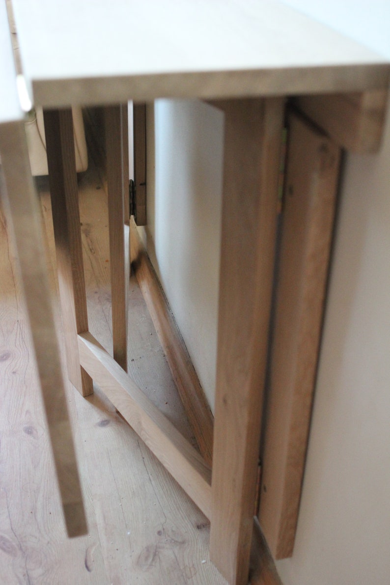 May include: A wooden folding table with a white top and a natural wood finish. The table is folded up and leaning against a wall.