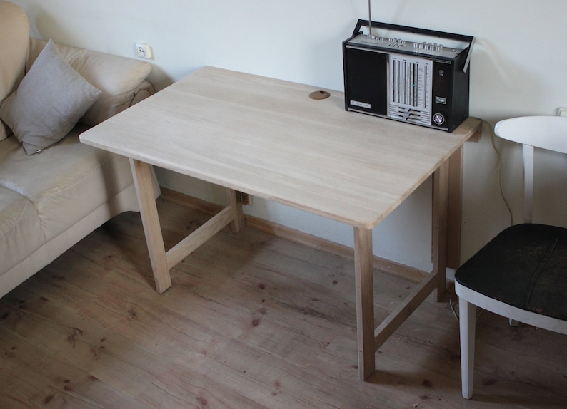 May include: A light wood table with a simple design, featuring a rectangular top and four legs. The table is in a room with a white couch and a black radio on top.