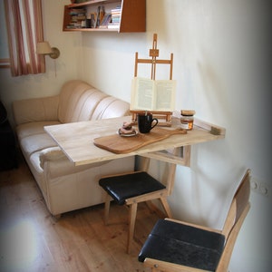 Puede incluir: Una mesa plegable de madera clara con una tabla de cortar de madera en la parte superior. La mesa est&aacute; unida a la pared y tiene dos sillas negras con patas de madera. Hay un libro en un caballete y una taza en la mesa.