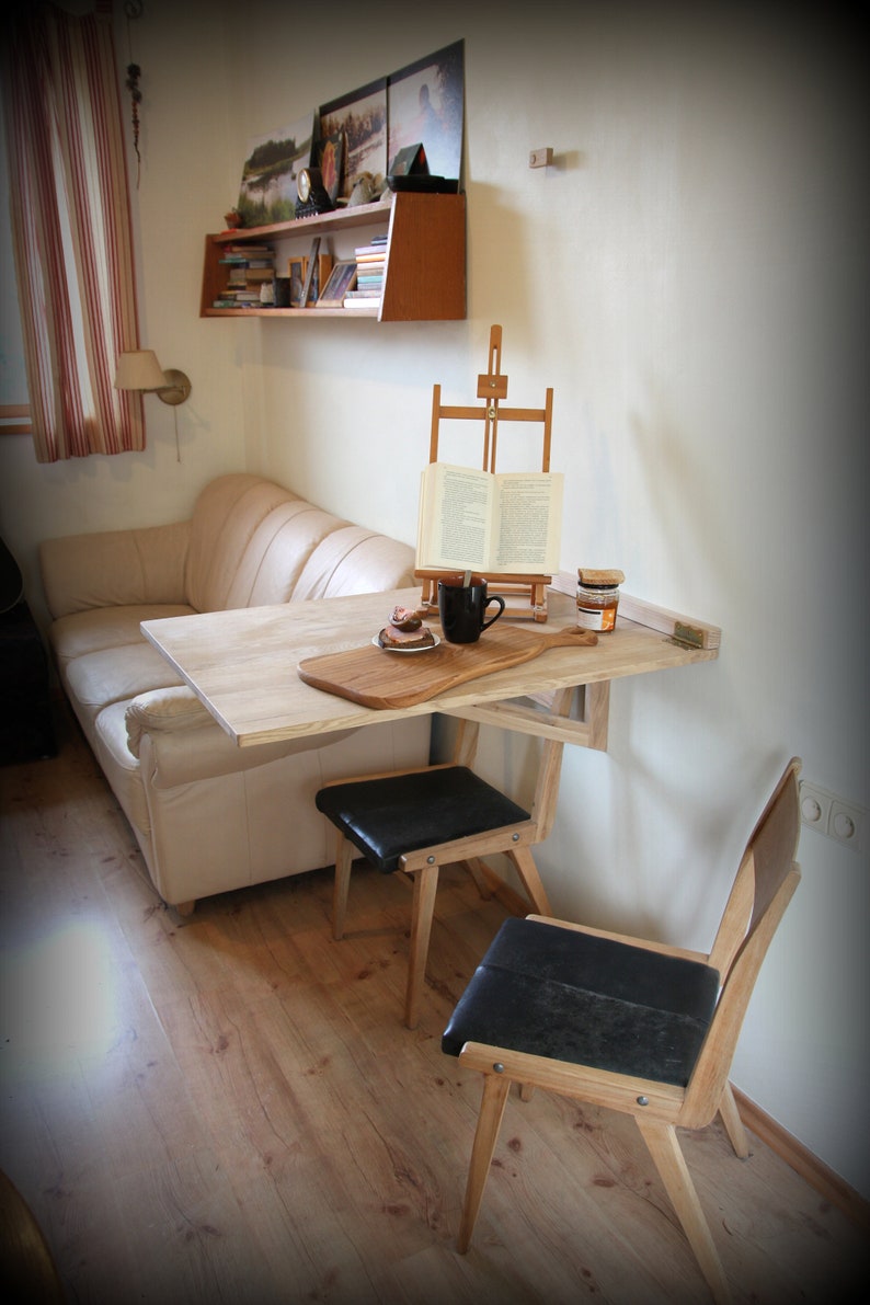 Puede incluir: Una mesa plegable de madera clara con una tabla de cortar de madera en la parte superior. La mesa est&aacute; unida a la pared y tiene dos sillas negras con patas de madera. Hay un libro en un caballete y una taza en la mesa.