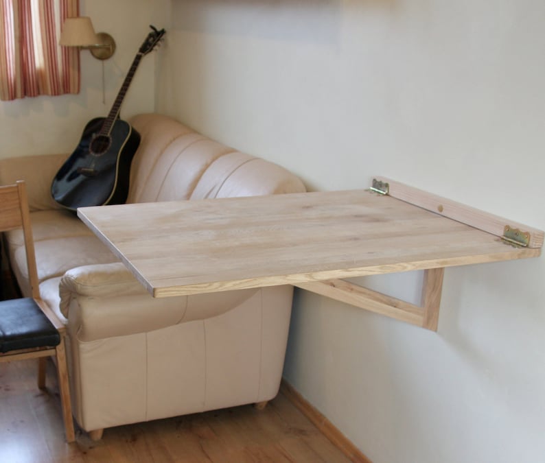 Puede incluir: Una mesa plegable de madera clara montada en una pared con bisagras. La mesa est&aacute; plegada y est&aacute; unida a una pared junto a un sof&aacute; de color claro.