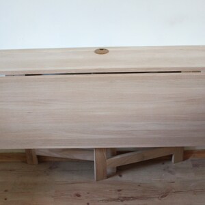 May include: A light wood folding table with a simple design. The table is closed and the legs are folded underneath. The table top is smooth and has a light wood grain.