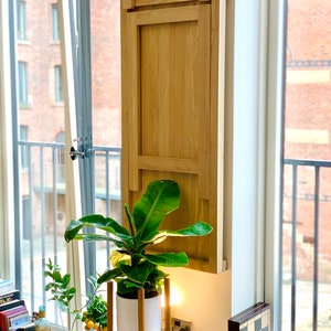 May include: A light wood folding table mounted to a wall, with a green plant in a white pot on a stand in front of it.