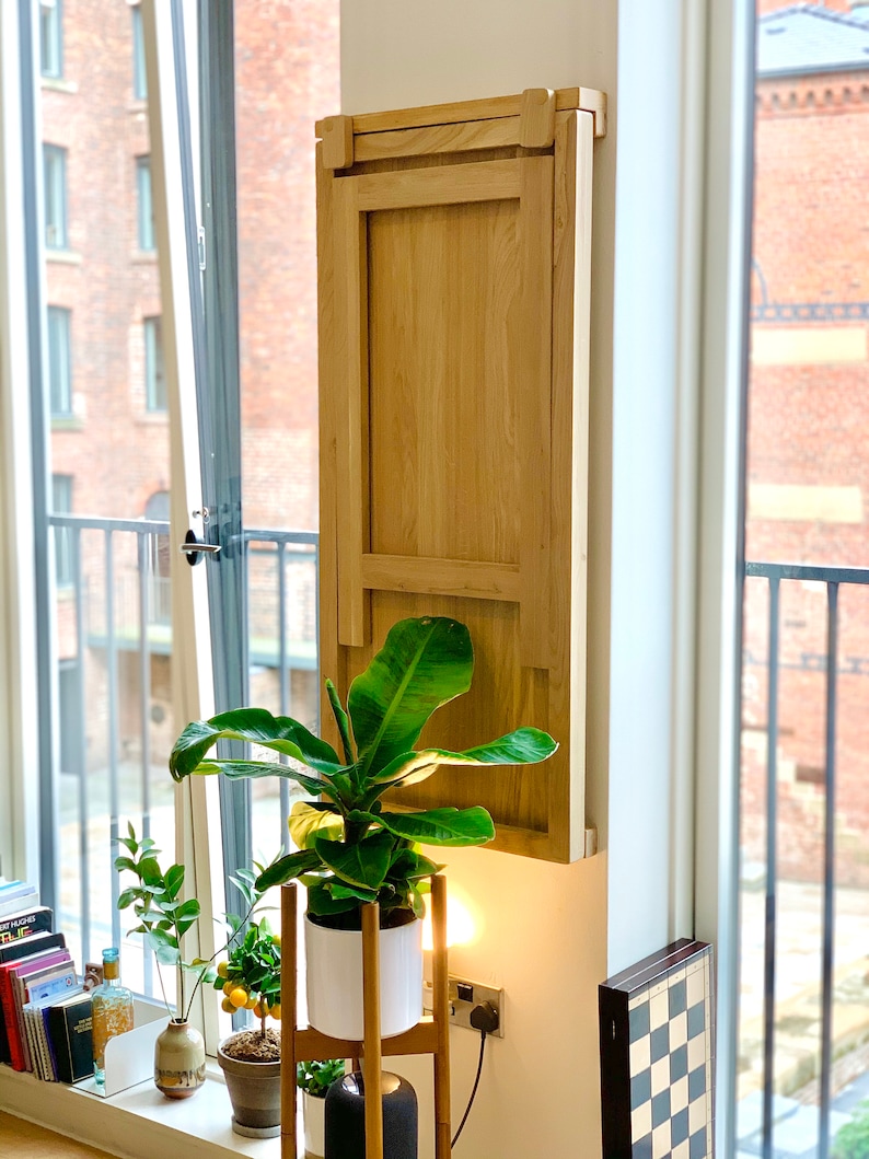 May include: A light wood folding table mounted to a wall, with a green plant in a white pot on a stand in front of it.