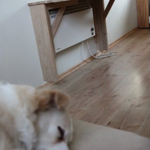 May include: A white electric heater mounted on a wall with a wooden folding table above it. The table is made of light wood and has a simple design. The heater has a white finish and a black knob. The table is folded up and is not in use.