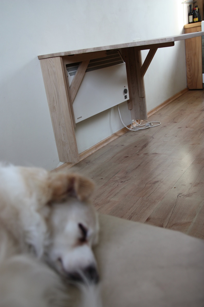 May include: A white electric heater mounted on a wall with a wooden folding table above it. The table is made of light wood and has a simple design. The heater has a white finish and a black knob. The table is folded up and is not in use.