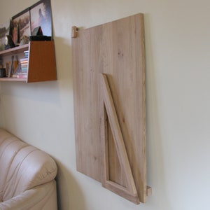 Puede incluir: Una mesa plegable de madera clara montada en una pared. La mesa est&aacute; plegada y asegurada con un soporte triangular.