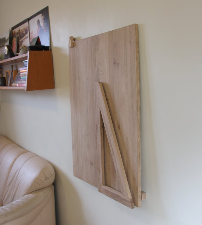 Puede incluir: Una mesa plegable de madera clara montada en una pared. La mesa est&aacute; plegada y asegurada con un soporte triangular.