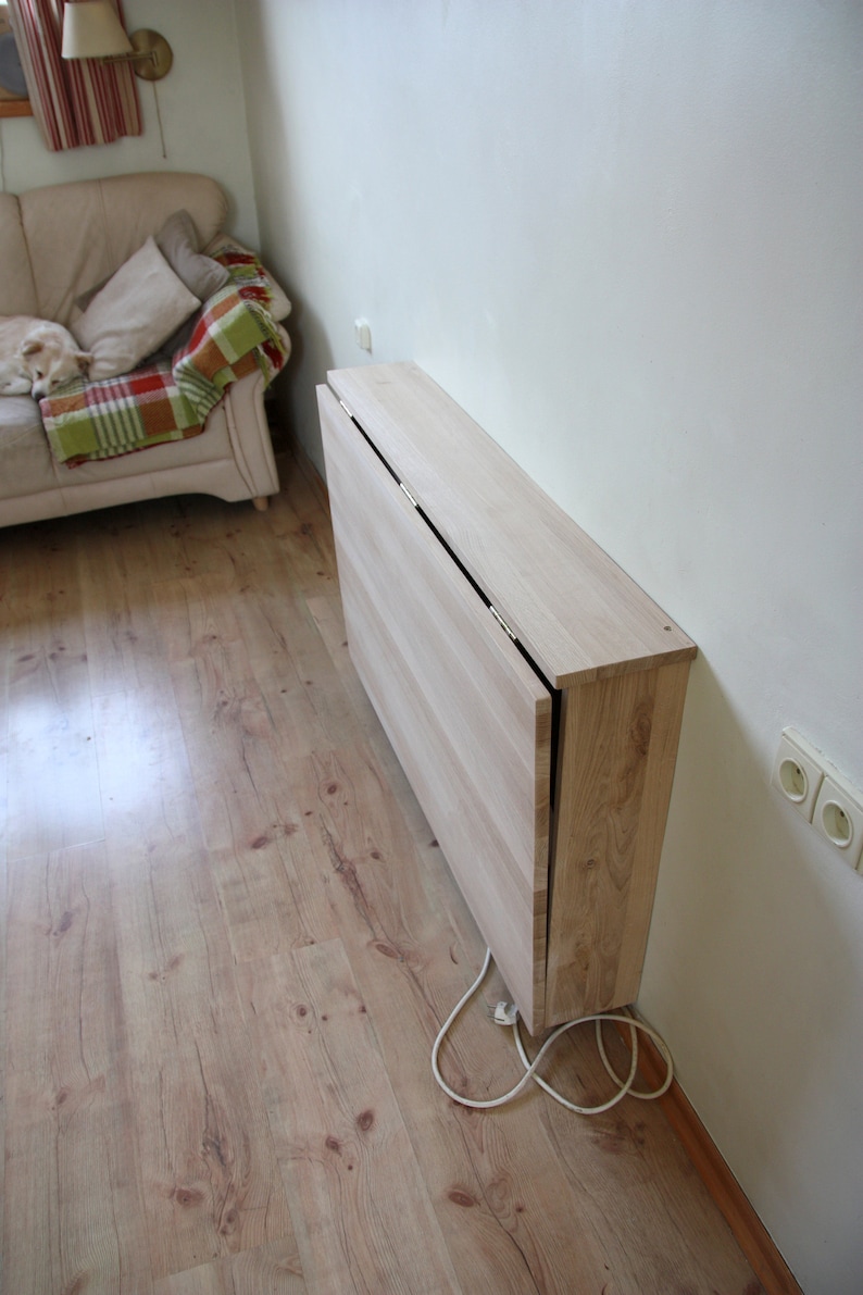 May include: A light wood folding table mounted to a wall. The table is closed and folded up against the wall. The table is in a room with light wood floors.