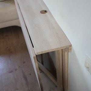 May include: A white wooden folding table mounted to a wall. The table is folded up and against the wall, with the top surface facing the camera. The table is made of light wood and has a simple design.