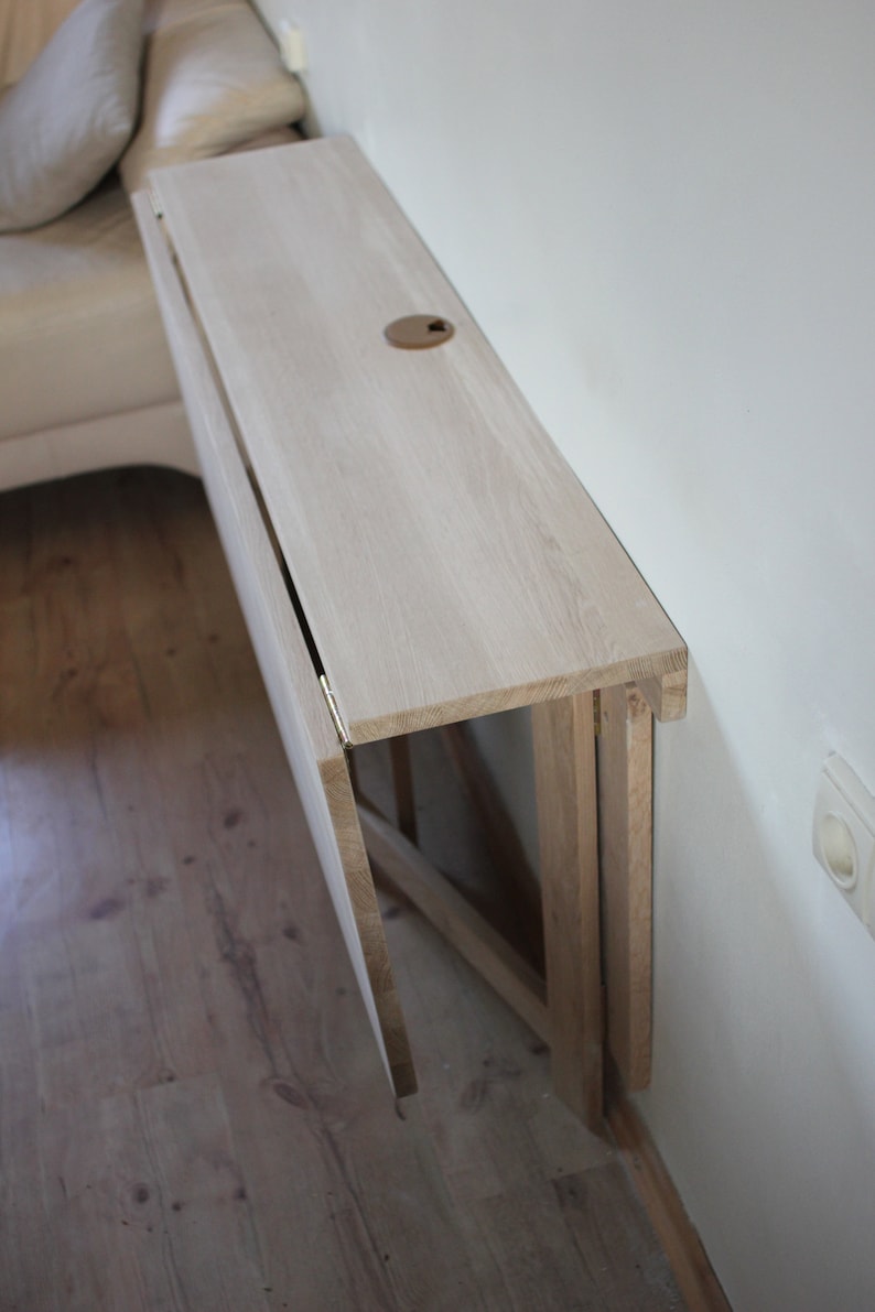 May include: A white wooden folding table mounted to a wall. The table is folded up and against the wall, with the top surface facing the camera. The table is made of light wood and has a simple design.