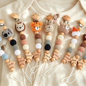 May include: A collection of baby pacifier clips with wooden beads and crochet animal toppers. Each clip features a different animal, including a koala, lion, bear, fox, and rabbit. The clips are personalized with names on wooden star-shaped charms.