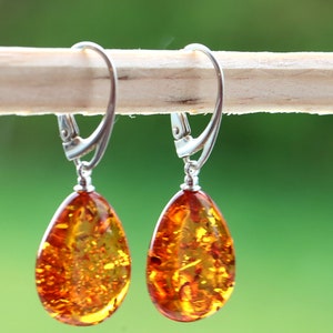 May include: A pair of amber teardrop earrings with silver-colored leverback closures. The amber stones are a warm, translucent orange-brown color, with visible internal patterns. The earrings are displayed against a blurred green background.
