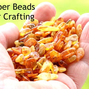 May include: A collection of small, polished, translucent amber beads in various shades of yellow, orange, and brown. The text "Amber Beads For Crafting" is printed in black in the upper left corner of the image.
