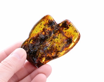 May include: A polished, irregular piece of translucent amber featuring deep golden-yellow hues and dark, dense organic inclusions throughout its structure. The stone has a smooth, rounded surface with a glossy finish, showcasing natural variations in color.