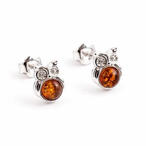 May include: A pair of silver stud earrings shaped like owls. Each owl features a round, polished amber stone as the body and two small, clear, sparkling stones set as the eyes. The earrings include silver post backs for secure attachment.