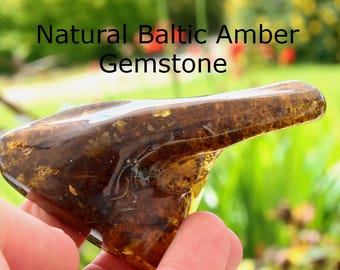 May include: A polished, irregular piece of dark brown, translucent Baltic amber gemstone with visible internal inclusions and organic textures. The text Natural Baltic Amber Gemstone is printed in black letters above the stone.