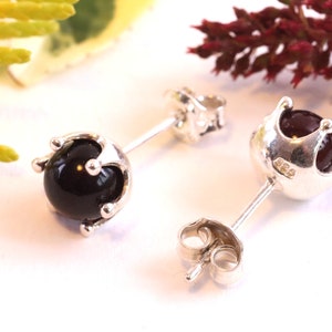 Könnte beinhalten: Ein Paar silberne Ohrstecker mit schwarzen und roten runden Edelsteinen, die in einem kronenartigen Design gefasst sind.